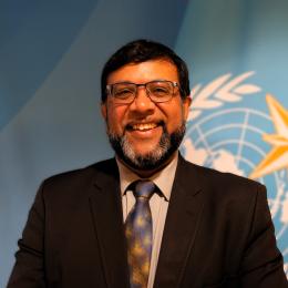 A man in a suit and tie smiles in front of a blue background featuring a United Nations-style emblem with a globe, laurel branches, and a star.