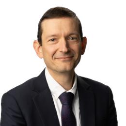 Man in a dark suit and tie, smiling slightly, posing against a plain white background.