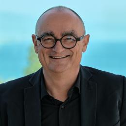 A smiling man with glasses, short hair, and a black jacket stands outdoors with a blurred blue background.