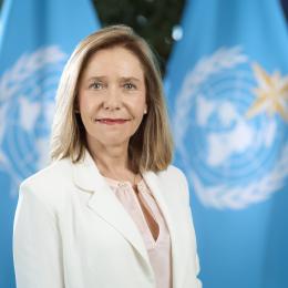 A woman in a white blazer stands in front of two blue flags featuring the United Nations emblem.