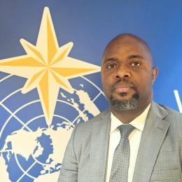 A man in a gray suit and tie stands in front of a blue wall featuring a yellow star and a world map design.