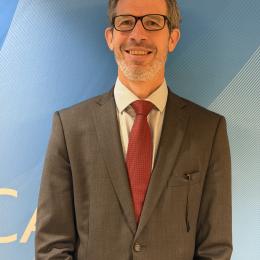 A man wearing glasses, a gray suit, white shirt, and red tie stands in front of a blue patterned background, smiling at the camera.