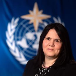 A woman with long dark hair in a black suit stands in front of a blue backdrop featuring a white emblem with a globe, laurel wreath, and a gold star.
