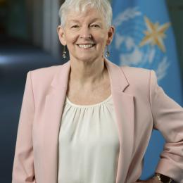 An older woman with short white hair, wearing a light pink blazer and cream top, stands smiling with one hand on her hip in front of a blue flag with a yellow star.