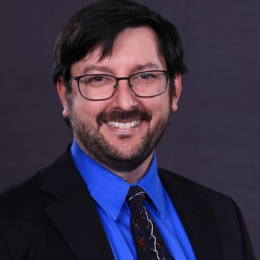 Man with dark hair, glasses, and beard wearing a blue shirt, patterned tie, and dark suit jacket, smiling in front of a plain dark background.