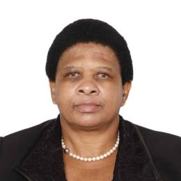 A woman with short black hair wears a black jacket, a lacy black top, and a pearl necklace, facing forward against a plain white background.