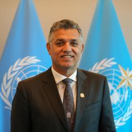 A man in a suit and tie standing in front of two United Nations flags.