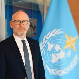 A man in a suit and glasses standing next to a blue World Meteorological Organization flag.