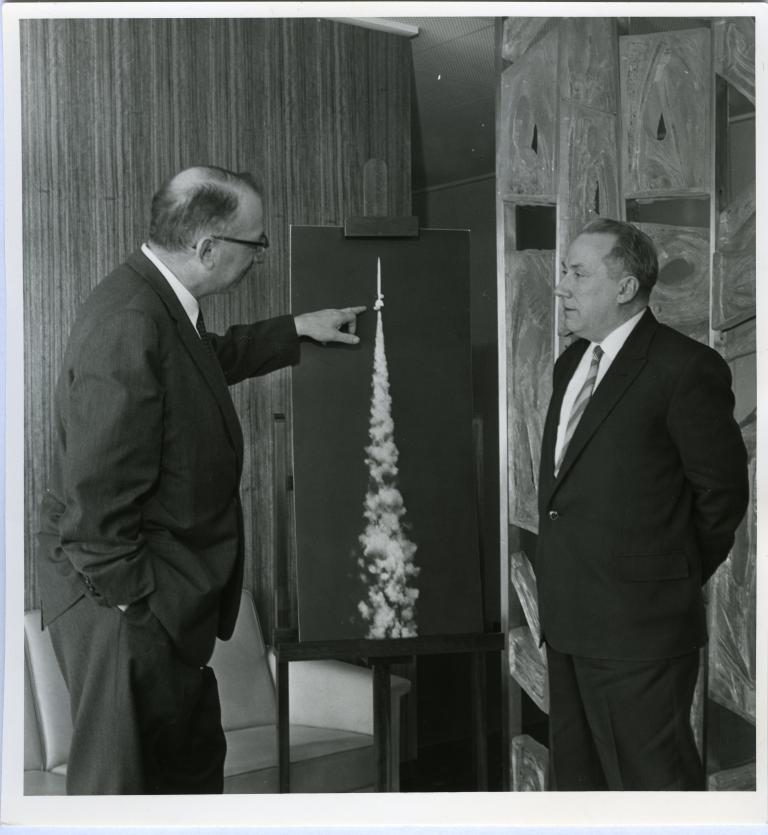 Two men standing in front of a painting of a rocket.