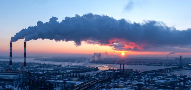 The sun is setting over a factory with smoke coming out of the chimneys.