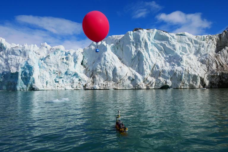A large red balloon hovers above a small boat near an iceberg in a calm sea under a clear sky.