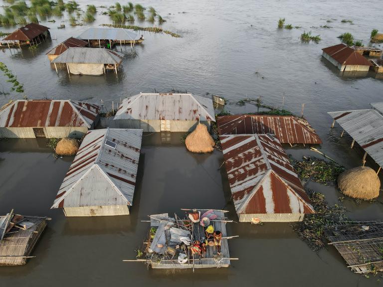 Houses partially submerged in floodwaters with individuals standing on a raft-like structure amidst the inundated area.