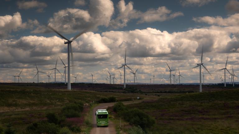 A bus travels along a rural road amidst a vast wind farm under a cloudy sky.