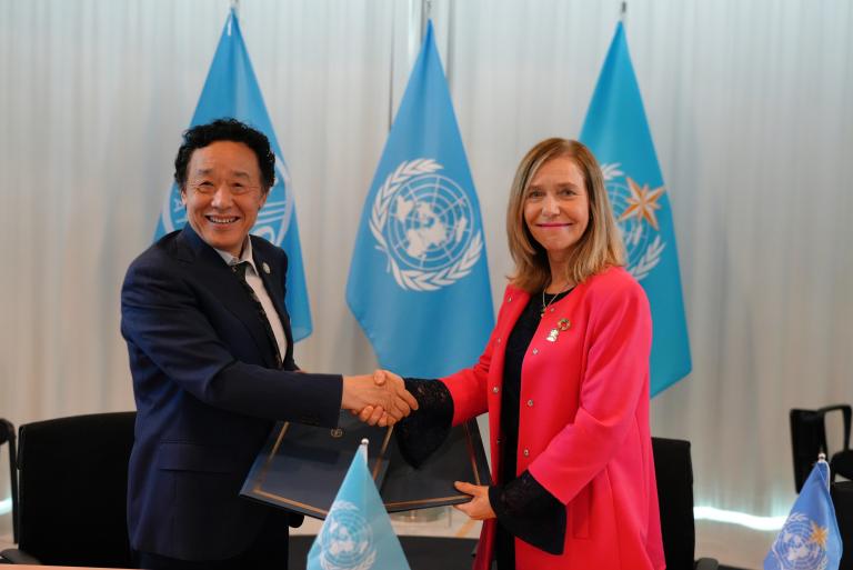 Two individuals, one in a dark suit and the other in a red jacket, are shaking hands with blue United Nations flags in the background.