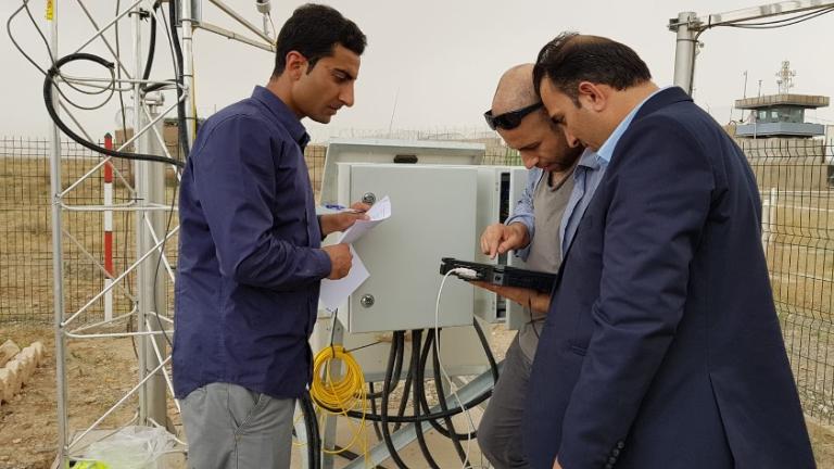 Three men stand outdoors examining equipment near a metal box with cables. One holds papers, another operates a device, and a third observes. A metal tower and fenced area are in the background.