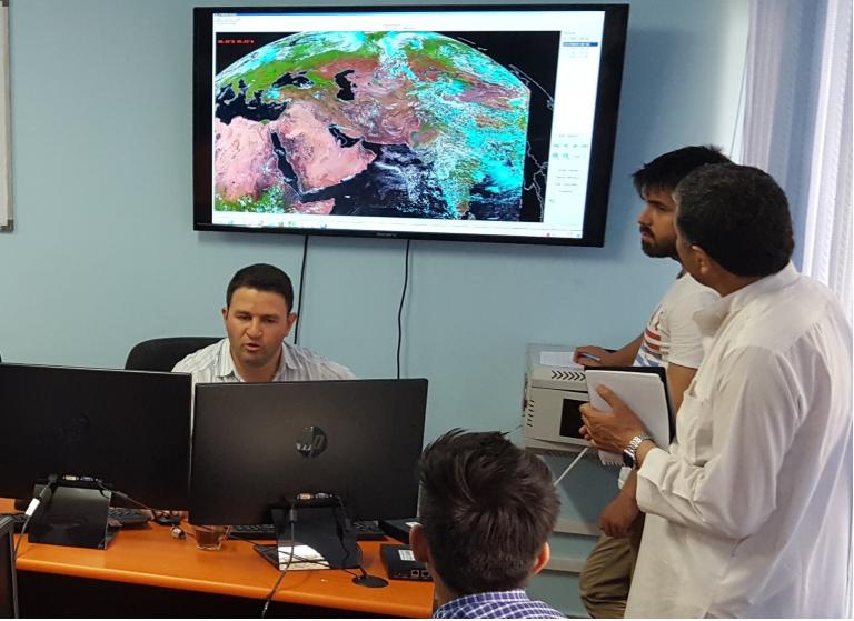 Four men are in a room with three computer screens, analyzing satellite imagery displayed on a large monitor. One man is seated at a desk, while the others are standing and holding documents.