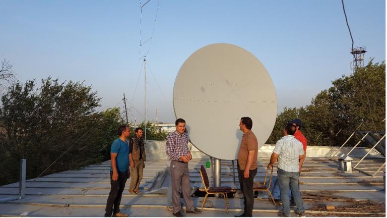 A group of five men stand on a rooftop, surrounding a large satellite dish. The rooftop has trees and structures in the background.