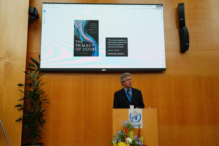 A man stands at a podium with a bouquet of flowers, speaking in front of a large screen displaying the cover of the book 