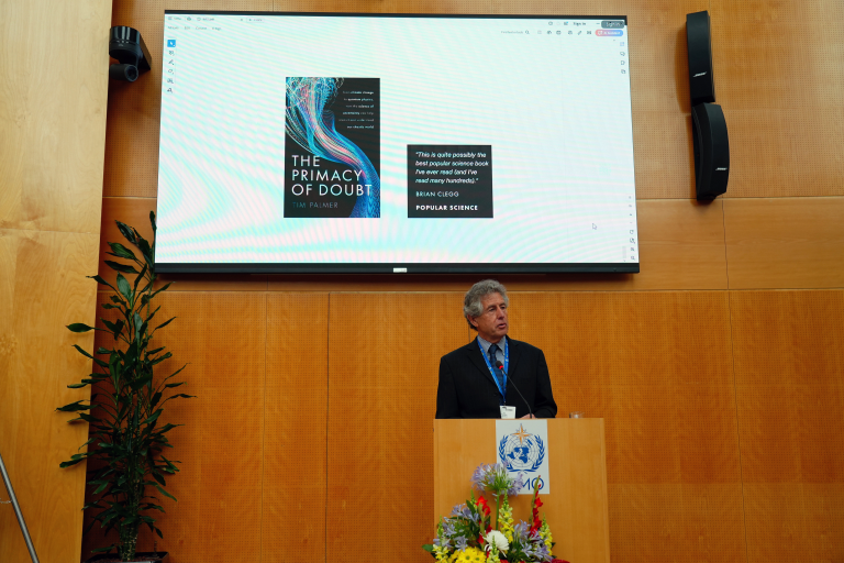 A man is giving a presentation in a room with wood-paneled walls. A large screen behind him displays the book "The Primacy of Doubt" by Tim Palmer with a quote praising the book.