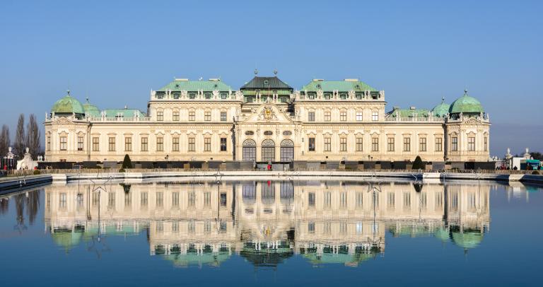 A large baroque-style palace with multiple green-roofed domes and intricate details, reflected in a still pond in front.