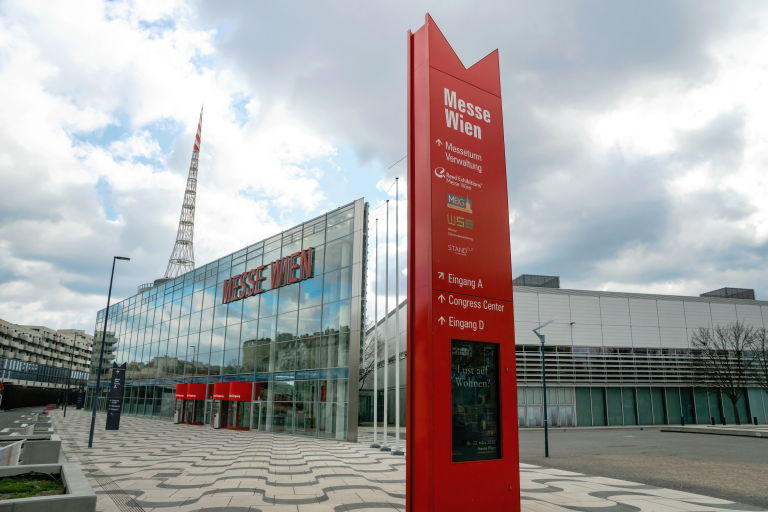 The image shows Messe Wien, a convention and exhibition center in Vienna, Austria. A tall red sign with directions stands in front of the modern glass building under a cloudy sky.