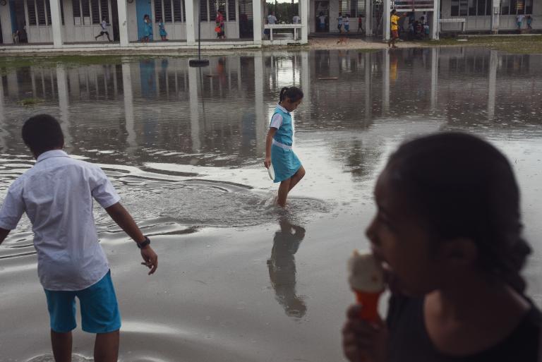 Children walk through a flooded schoolyard after rain, while one child in focus is eating an ice cream in the foreground.