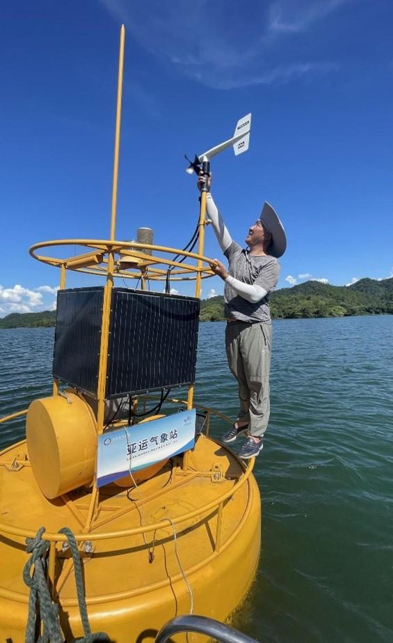 A person stands on a yellow buoy in the water, adjusting a weather instrument. The buoy features solar panels and a sign with text. The sky is clear, and land with greenery is visible in the background.