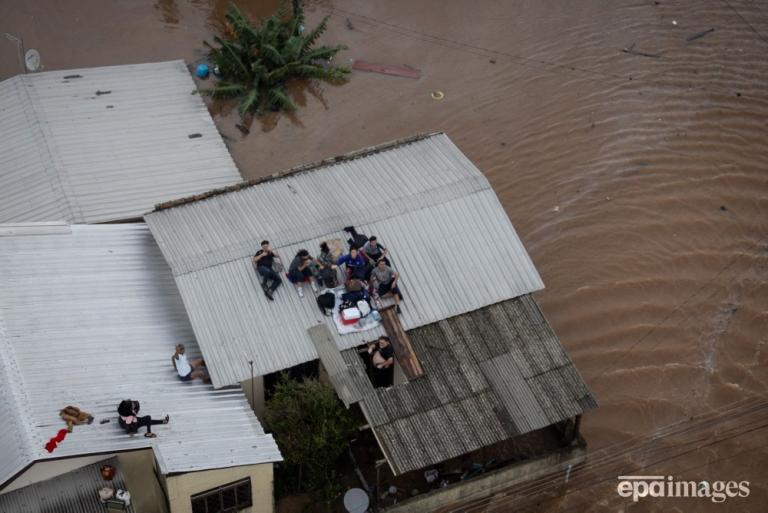 Aerial view in Brazil of stranded people on rooftops surrounded by floodwater, with partially submerged houses and visible debris in the water.