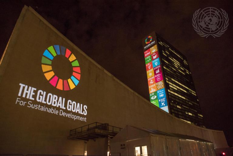 Night view of an illuminated UN building with "The Global Goals for Sustainable Development" logo projected on the side, while colorful Sustainable Development Goals icons display on an adjacent tower.