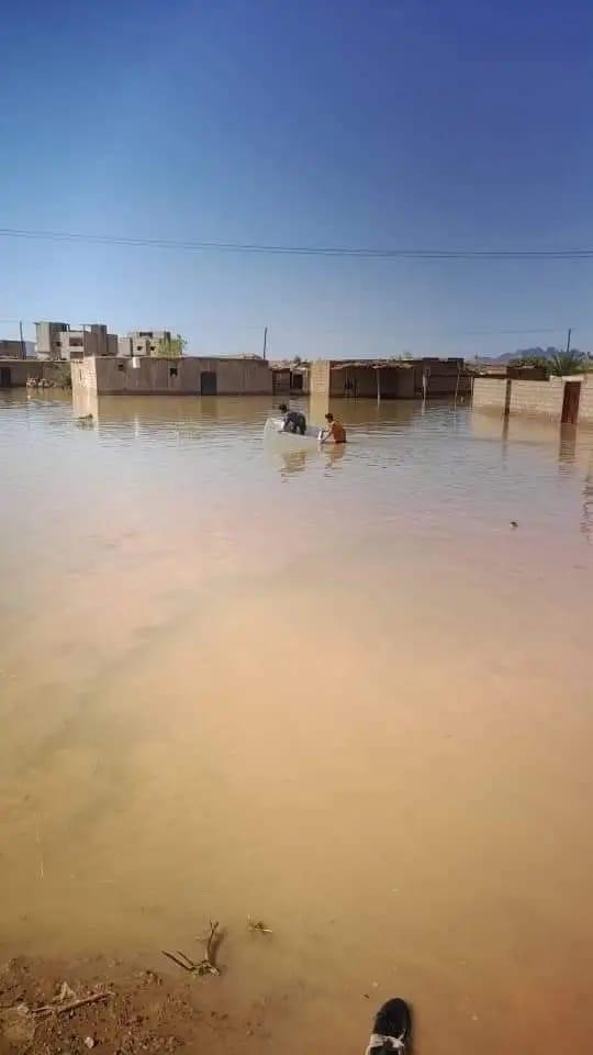 Two people navigate a small boat through a flooded area with partially submerged buildings in the background under a clear sky.