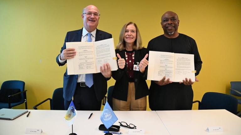 Three individuals stand in front of a table, holding up documents. The person in the middle is giving two thumbs up. An United Nations flag is visible on the table.