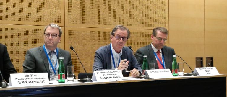 Three men in suits sit at a conference table with microphones and nameplates. The man in the middle speaks, while the others listen attentively. Water bottles are on the table.