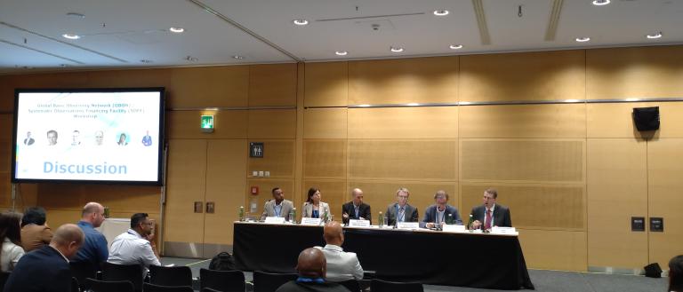 A panel of six people sits behind a table on a stage during a discussion, with an audience seated in front of them. A screen on the left displays the text "Discussion.