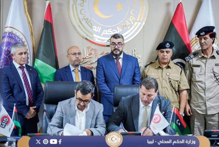 Officials gather around a table as two men sign documents, with flags and an emblem in the background.