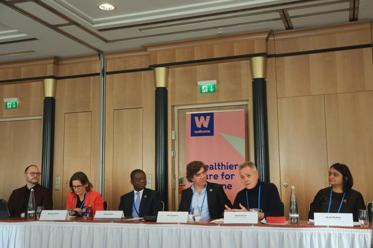 A panel of six people sits at a long table with microphones during a conference. A banner behind them displays the "Wellcome" organization name.