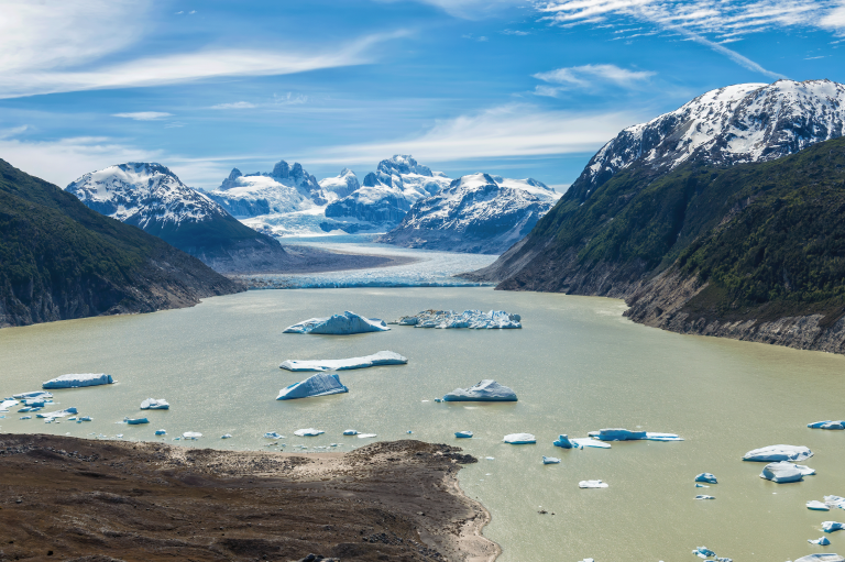 A glacier-fed lake with floating icebergs, bordered by rugged mountains and snow-capped peaks under a partly cloudy sky.