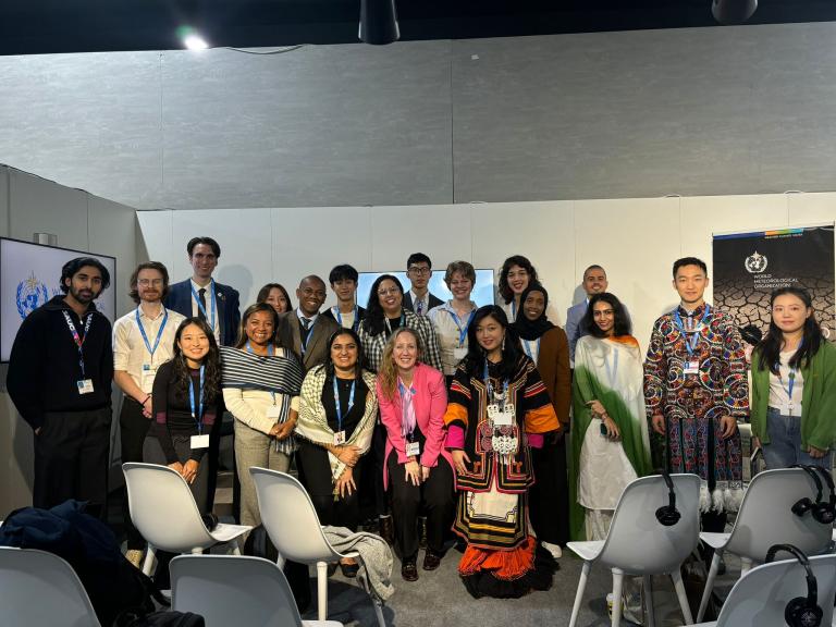 A diverse group of people pose for a photo in a conference room setting, with empty chairs in the foreground and banners in the background.