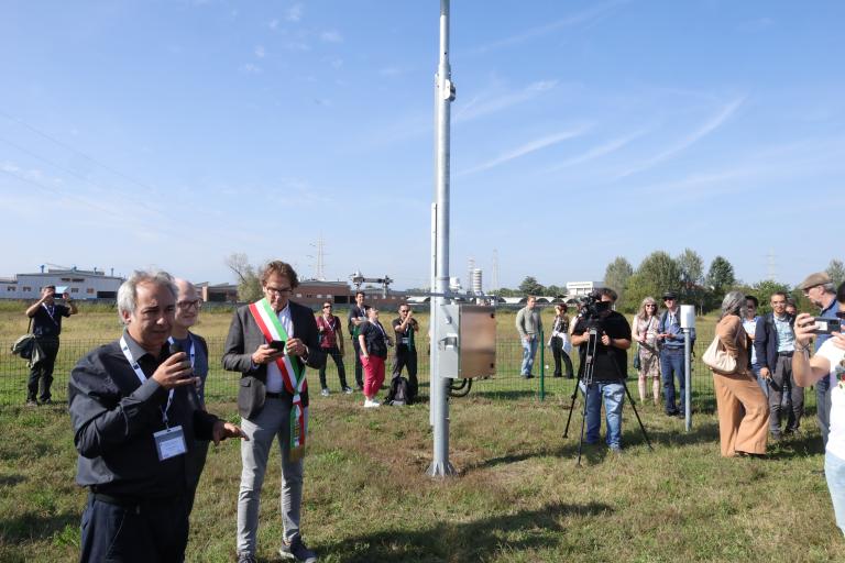 A group of people gather around a man speaking into a microphone in an open field with a pole-mounted device on a clear day.