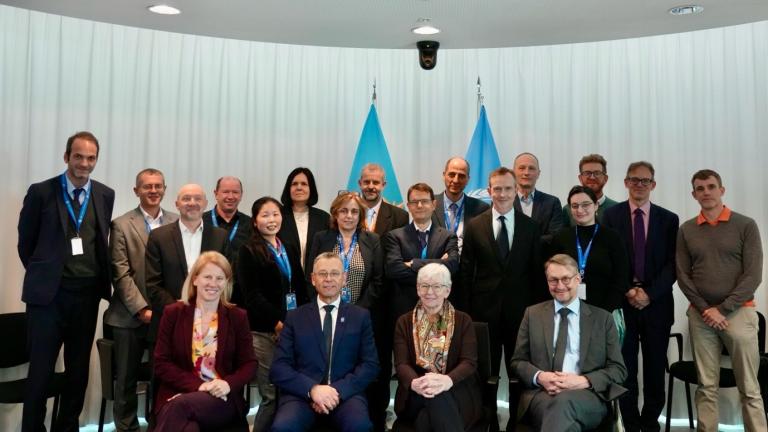 A group of twenty people in business attire pose for a photo in front of blue flags and a white curtain in a conference room.