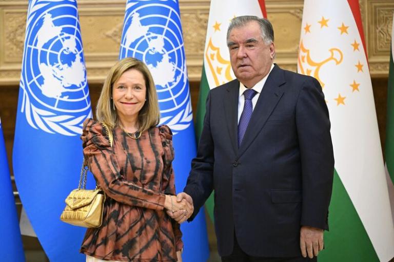 A woman and a man shake hands in front of United Nations and Tajikistan flags. Both are dressed formally and facing the camera.