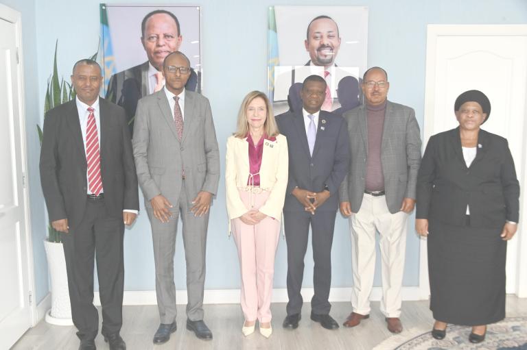 Six people pose for a group photo indoors, standing in a line with two framed portraits on the wall behind them.