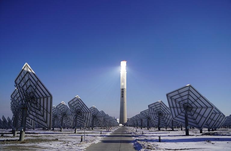 A solar power plant with multiple heliostat mirrors reflecting sunlight toward a central tower under a clear blue sky.