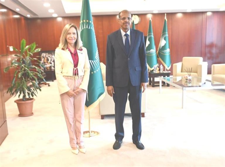 A woman and a man stand side by side in an office with wood-paneled walls, two green flags, a potted plant, and cream-colored furniture.