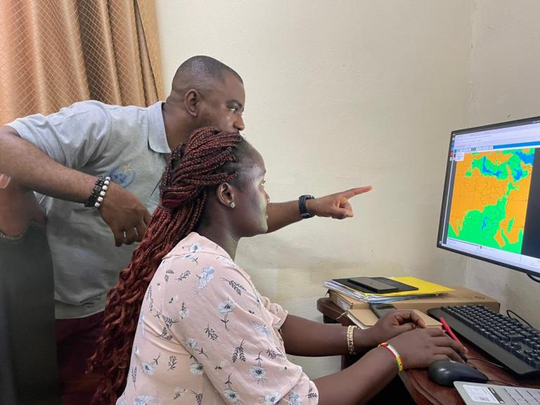 A man and a woman look at a computer screen displaying a colorful weather or geographic map in an office setting. The man points at the screen while the woman uses the mouse.