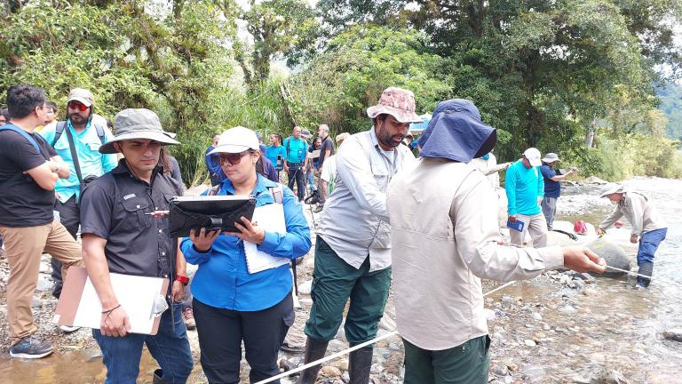 A group of people conduct field research beside a river, some taking notes, others using equipment and examining the water.