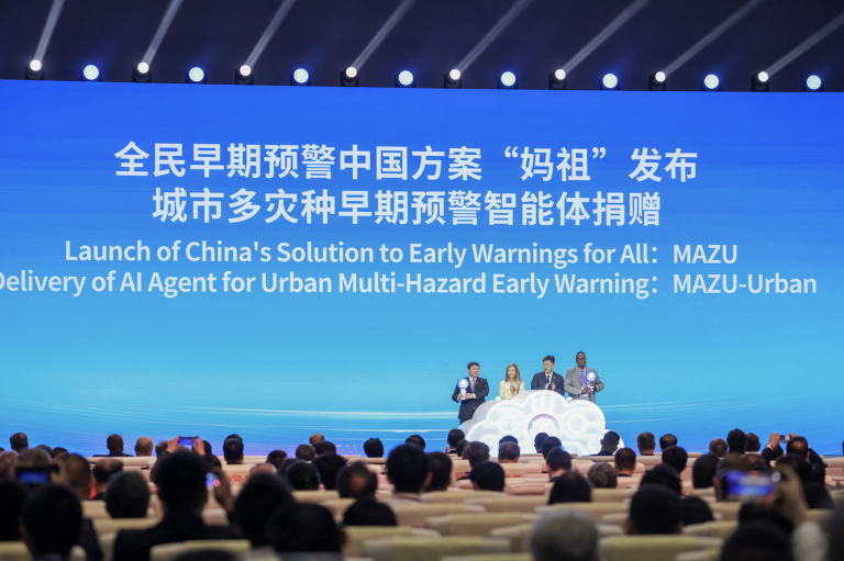 A large audience faces a stage where speakers stand in front of a blue screen displaying text about China's early warning AI solution, MAZU, for urban multi-hazard early warnings.