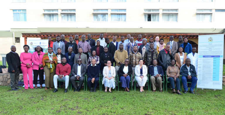 A large group of people pose for a photo outside a building, with informational banners on either side and a lawn in the foreground.