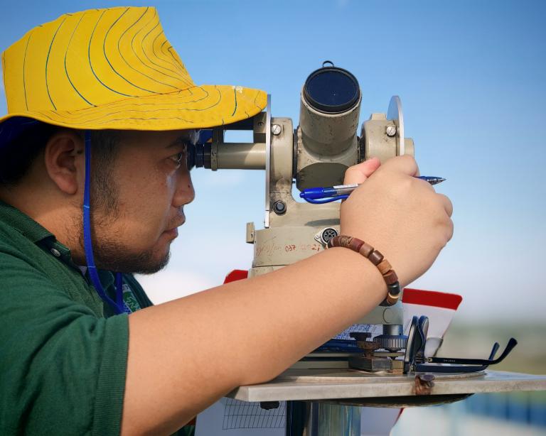 A person wearing a yellow hat looks through a surveying instrument while holding a pen and clipboard outdoors.