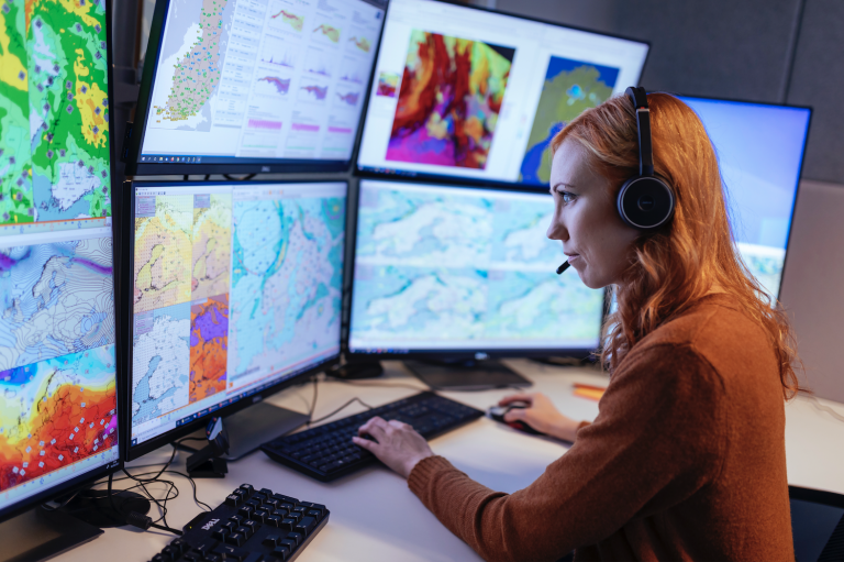 A woman wearing a headset analyzes weather maps and data on multiple computer monitors in an office setting.
