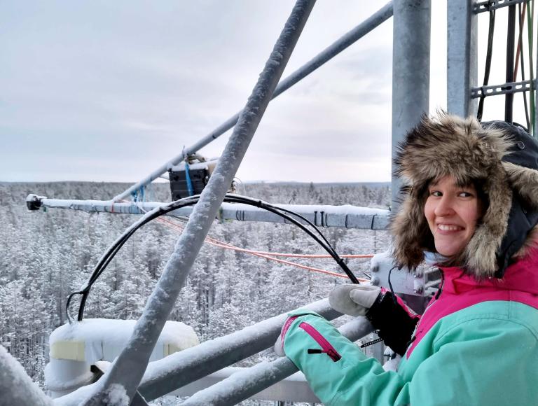 A person in a fur-lined hooded coat stands on a metal structure above a snowy forest, smiling at the camera.